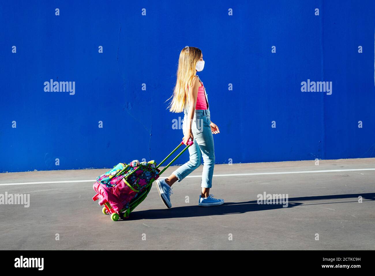 Blonde Mädchen zu Fuß mit Schultasche auf der Straße gegen blaue Wand Stockfoto