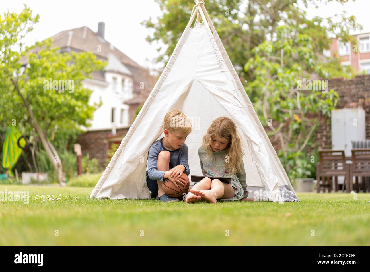 Geschwister mit digitalen Tablet sitzen im Zelt auf Gras an Hinterhof am Wochenende Stockfoto