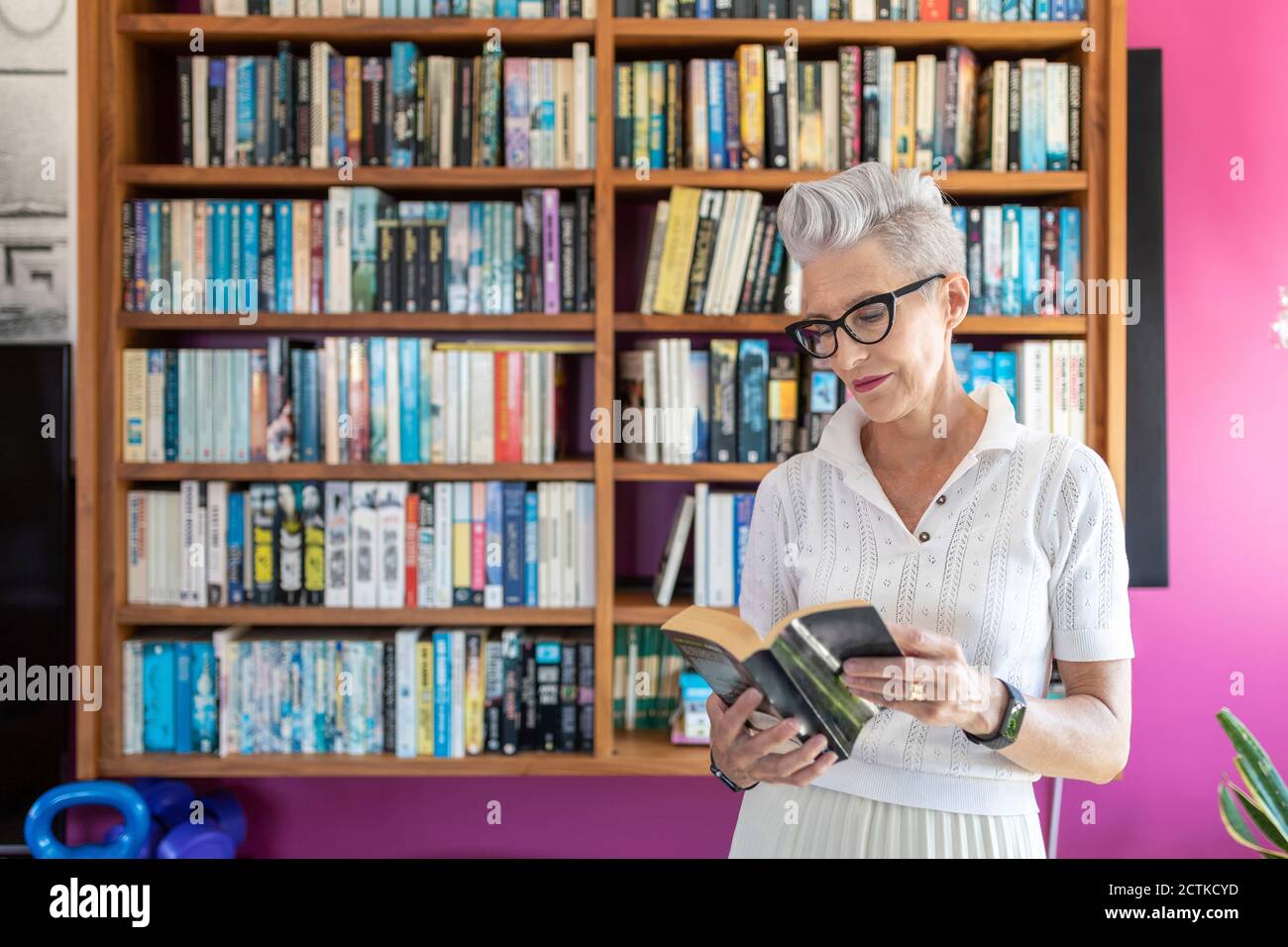 Aktive ältere Frau liest Buch, während sie gegen Bücherregal steht Zu Hause Stockfoto