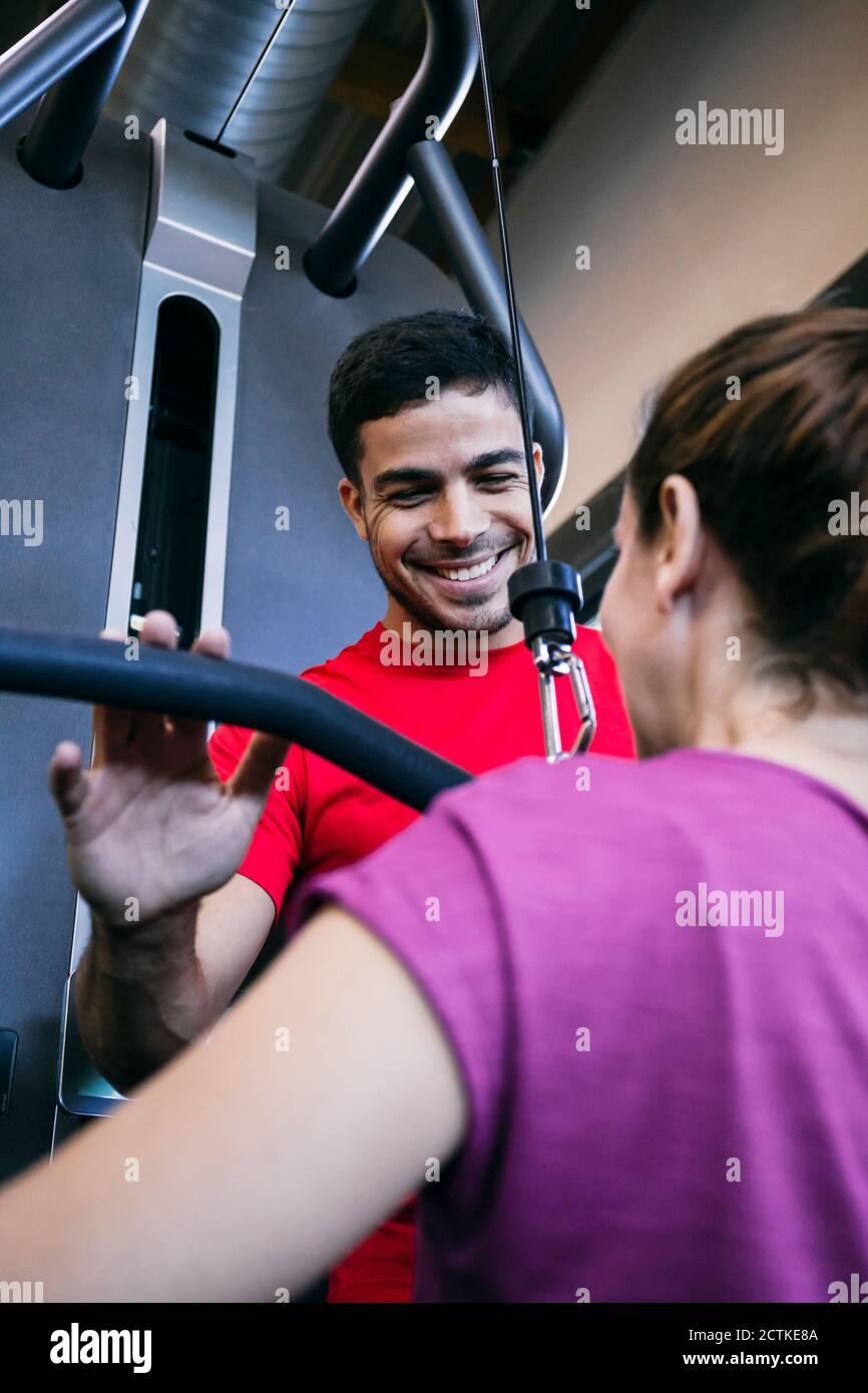 Lächelnder Fitnesstrainer unterstützt Frau beim Training mit seitlichem Pull-Down Gewichte Stockfoto