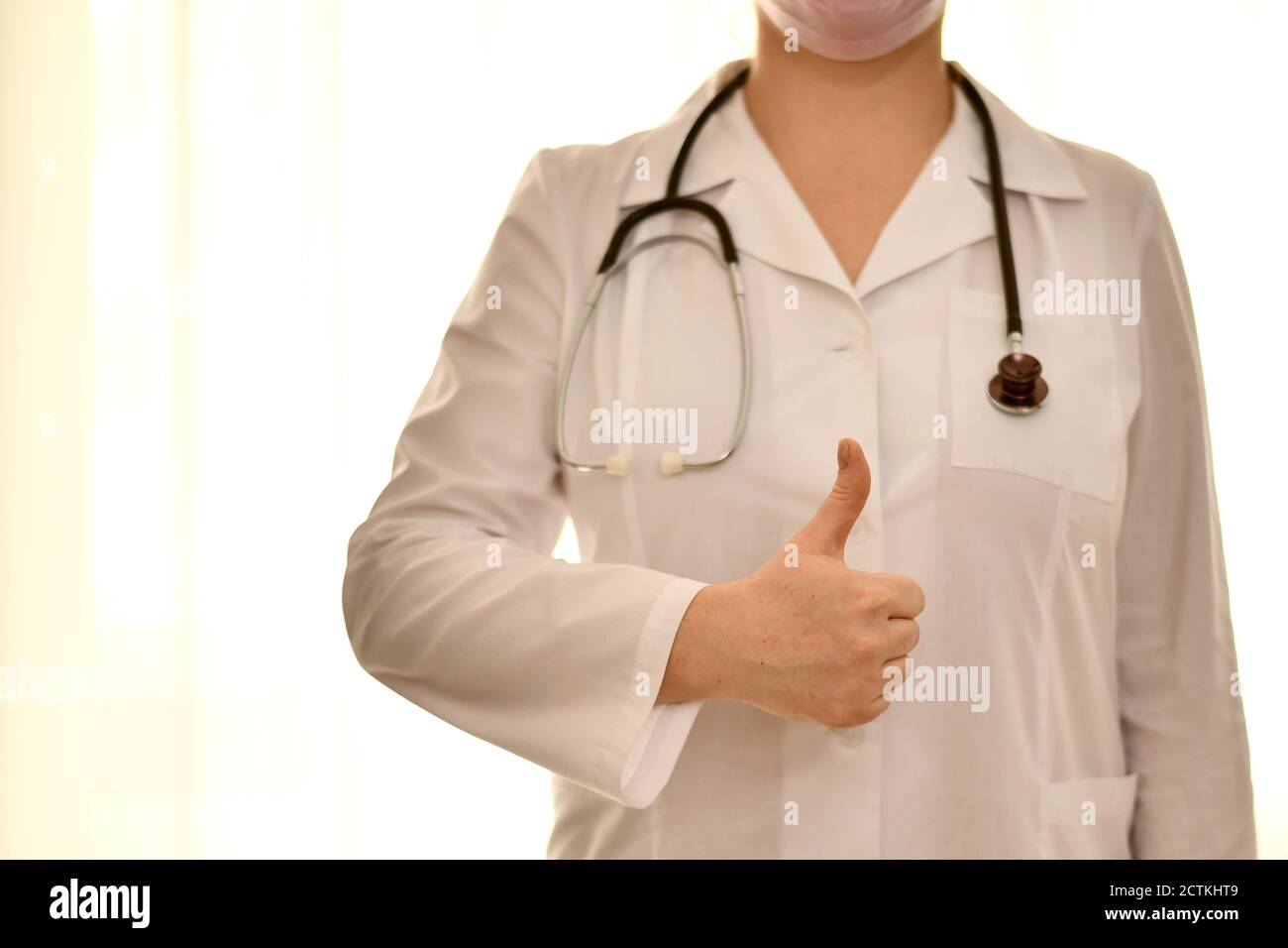 Arzt in weißer Robe mit Stethoskop zeigt Handgesten, ein gutes Symbol, einen erhobenen Daumen. Stockfoto