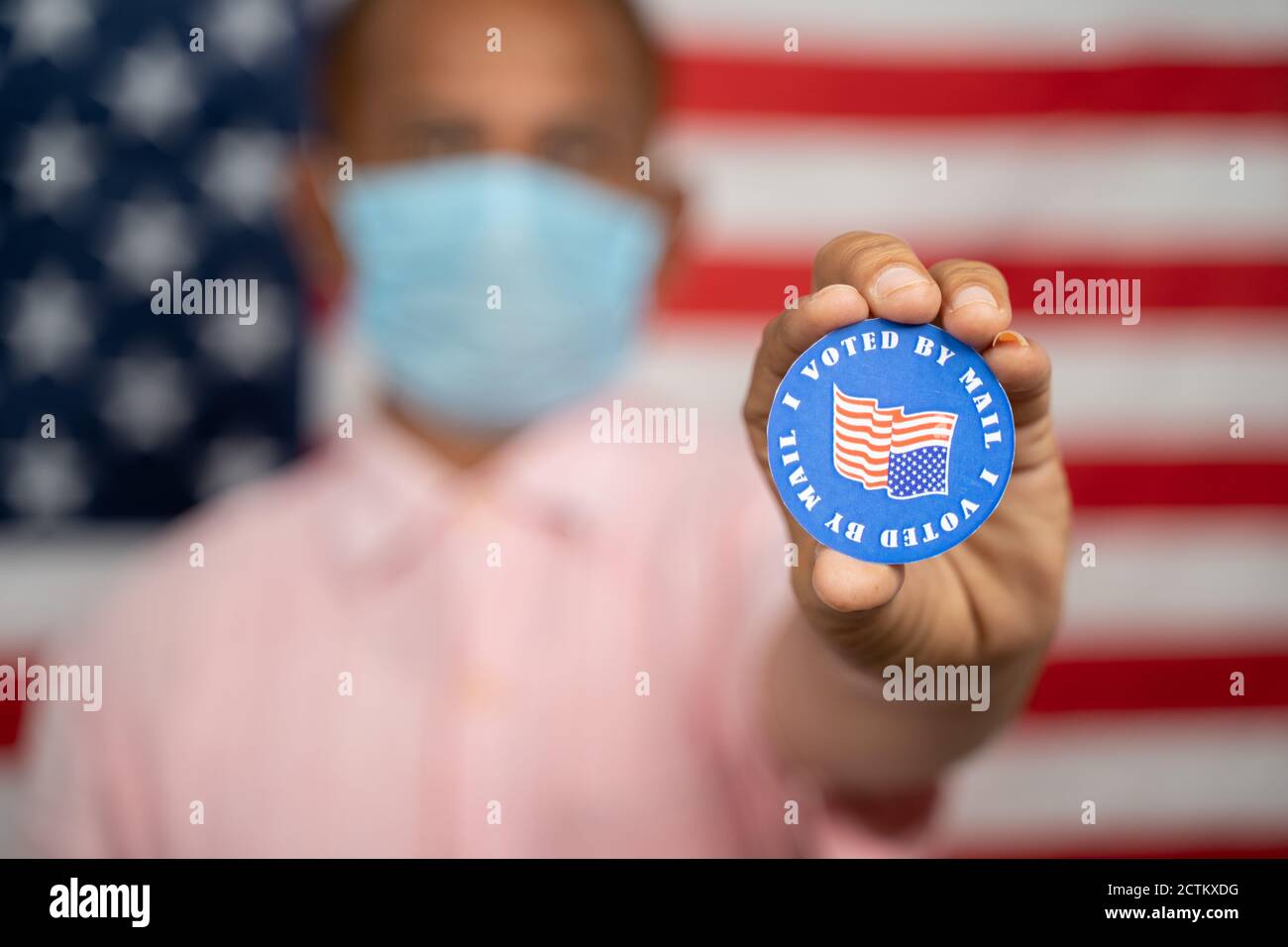 Mann in medizinischer Maske zeigt, dass ich per Mail-Aufkleber mit US-Flagge als Hintergrund gestimmt habe - Konzept der Post bei der Wahl in den USA. Stockfoto