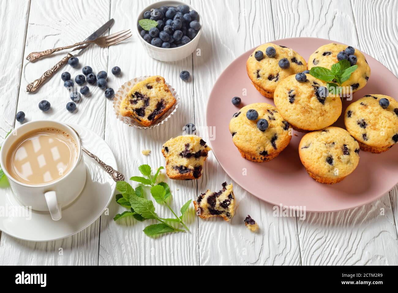 Heidelbeer Muffins gebacken mit frischen Heidelbeeren auf einem rosa Teller mit frischer Minze auf der Oberseite und Tasse Tee mit Milch auf weißem Holzhintergrund, Draufsicht, Stockfoto