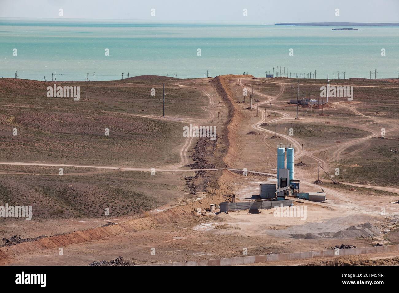 Mynaral/Kasachstan: Zementanlage von Jambyl. Einige Ausrüstung und Türme von Asphaltanlage. Weiße Straße auf Wüstenboden. Blick auf Balkhash Seeufer und Wüste. Stockfoto