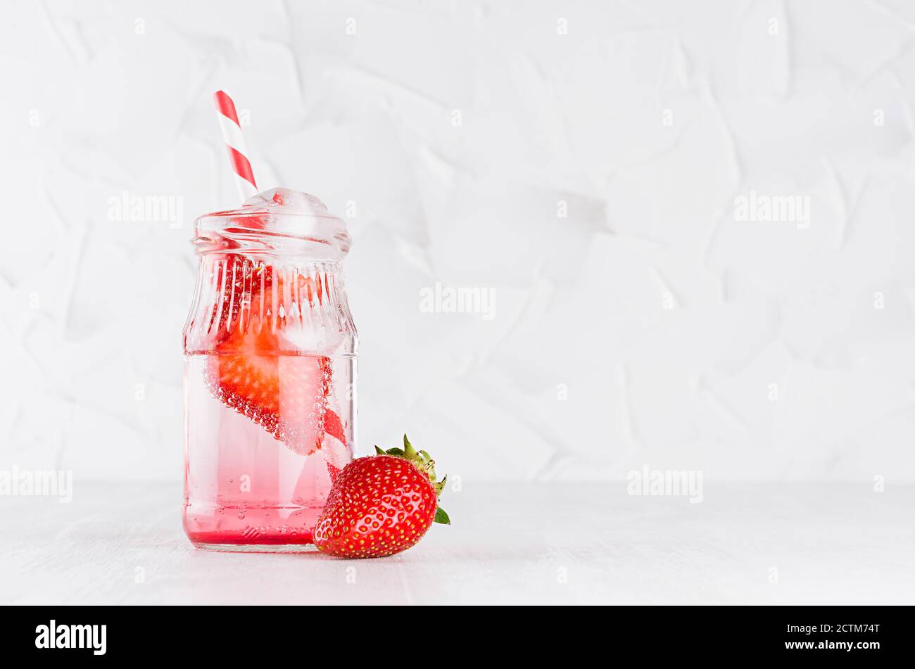 Erfrischendes kaltes Fruchtgetränk mit roter Erdbeere, Eis und Stroh im durchsichtigen Glas im eleganten weißen Interieur auf Holztisch, Kopierraum. Stockfoto