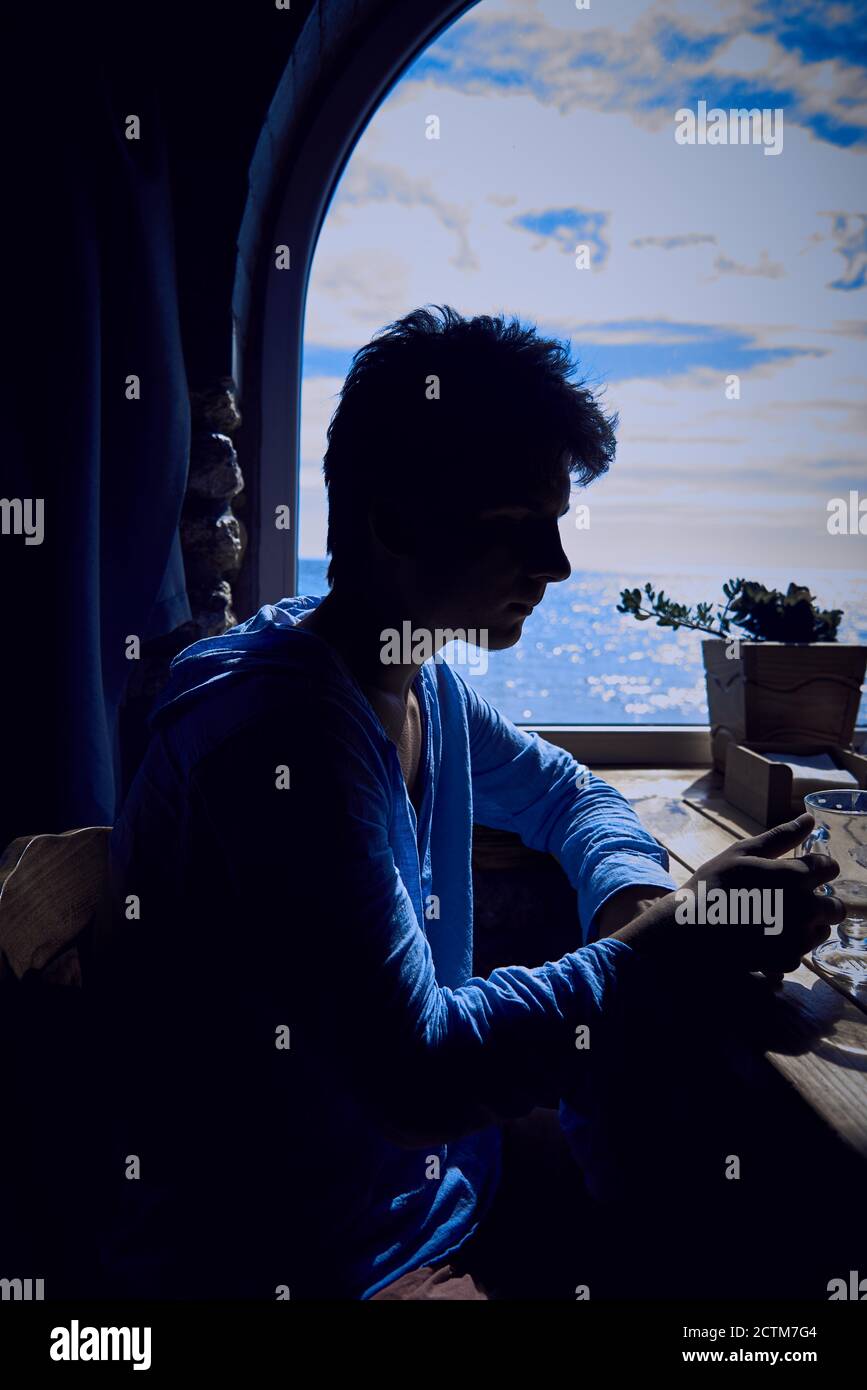 Silhouette eines jungen Mannes, der einen Cappuccino vor dem Hintergrund eines Fensters mit Meerblick beendet. Stockfoto