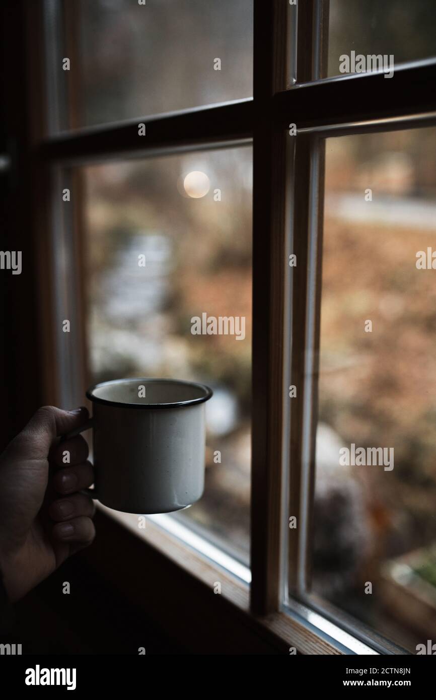 Crop unkenntlich Person mit Emaille Tasse heißen Kaffee stehen Fenster in der Nähe mit verschwommenem Blick auf die Natur an einem trüben Herbsttag Auf dem Land Stockfoto