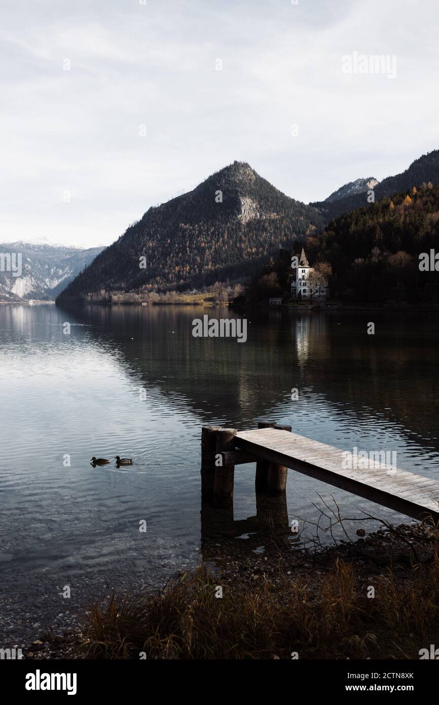 Herrliche Landschaft von ruhigen See mit ein paar Enten schwimmen In der Nähe von hölzernen Pier und einsamen Gebäude am Ufer gegenüber Felsige Berge im bewölkten Herbsttag Stockfoto