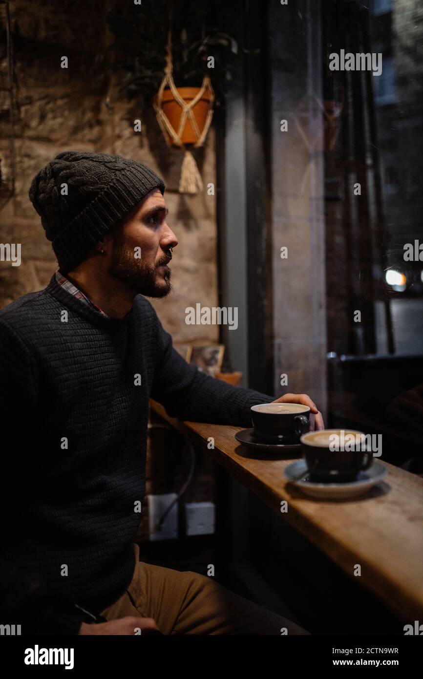 Seitenansicht des ruhigen Mannes in warmem Pullover und Hut Sitzen an der Theke in gemütlichen Holz-Café und genießen heiß Köstlicher Kaffee beim Blick weg Stockfoto