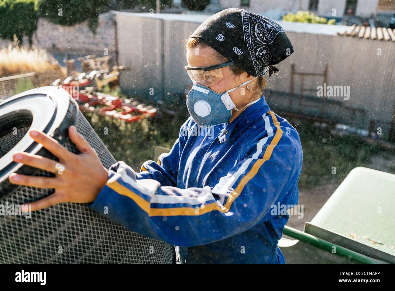 Seitenansicht der Bäuerin in Schutzbrille und Atemschutzmaske Maske Kontrolle und Reinigung Filter des landwirtschaftlichen Fahrzeugs während der Arbeit Auf dem Hof Stockfoto