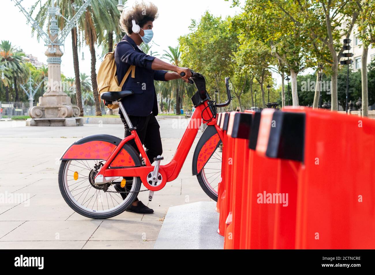 Junger schwarzer Mann in einem Anzug und Maske hören Musik mit dem öffentlichen Fahrrad auf der Straße Stockfoto