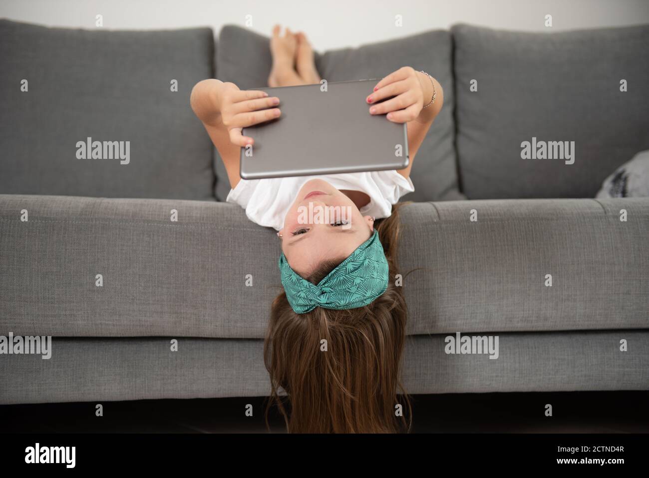 Kleines Mädchen liegt mit dem Gesicht nach unten auf der Couch, während sie unterhalten wird Verwendung von Tablet am Wochenende Stockfoto