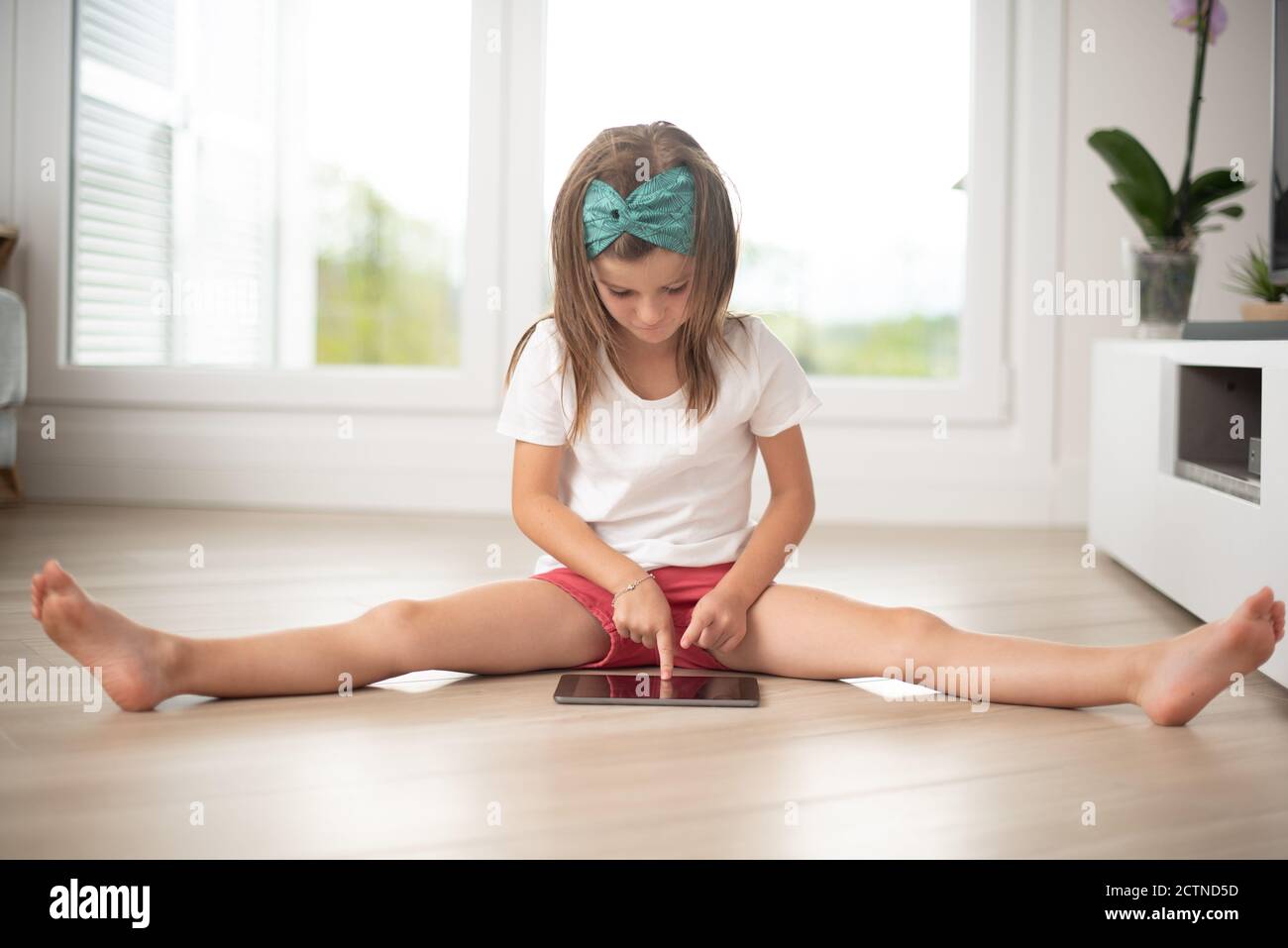 Kleines Mädchen auf dem Boden sitzen unterhalten mit Smartphone während Wochenende beim Stretching im Wohnzimmer Stockfoto