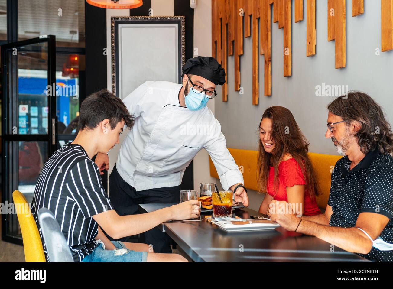 Männlich Koch trägt medizinische Maske Catering Gruppe von Inhalten Kunden Sitzen am Tisch im Café Stockfoto