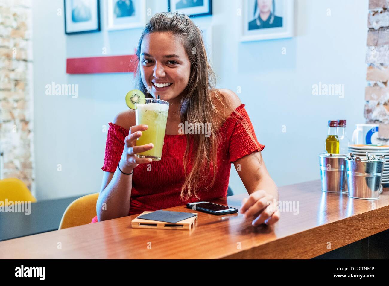 Lächelnde Kundin, die im Café am Tresen sitzt und genießt Süßes Detox-Getränk mit Kiwi beim Blick auf die Kamera Stockfoto