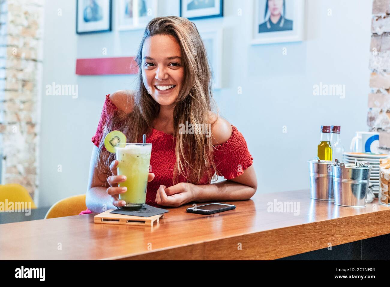 Lächelnde Kundin, die im Café am Tresen sitzt und genießt Süßes Detox-Getränk mit Kiwi beim Blick auf die Kamera Stockfoto