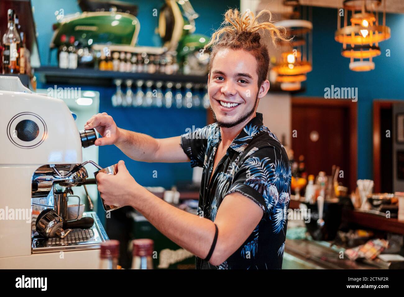 Seitenansicht des fröhlichen männlichen Barista mit Dreadlocks Vorbereitung aromatisch Kaffee in der Kaffeemaschine, während man die Kamera betrachtet Stockfoto