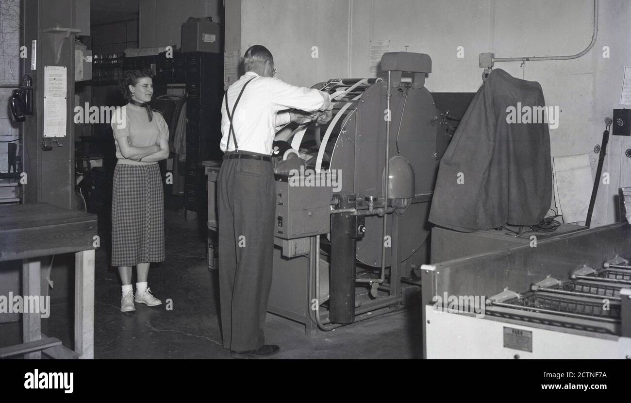 1940er Jahre, historisch, in einem Druckraum, eine Dame, die einem Ausflügler in Hosen und Hosenträger zusieht, und seine Jacke abschneidet, einen Streifen Papier auf einer Maschine ausschneidet, die Fotos von Filmnegativen, USA, druckte. Die Maschine war eine Kodak Papierverarbeitungsmaschine. Stockfoto