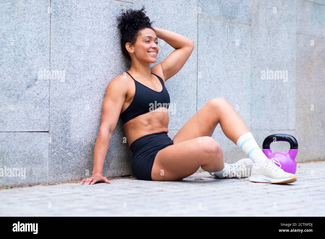 Positive schwarze athletische Frau in Sportkleidung sitzt auf Bürgersteig mit kettlebell und Pause beim Functional Workout in der Stadt Stockfoto
