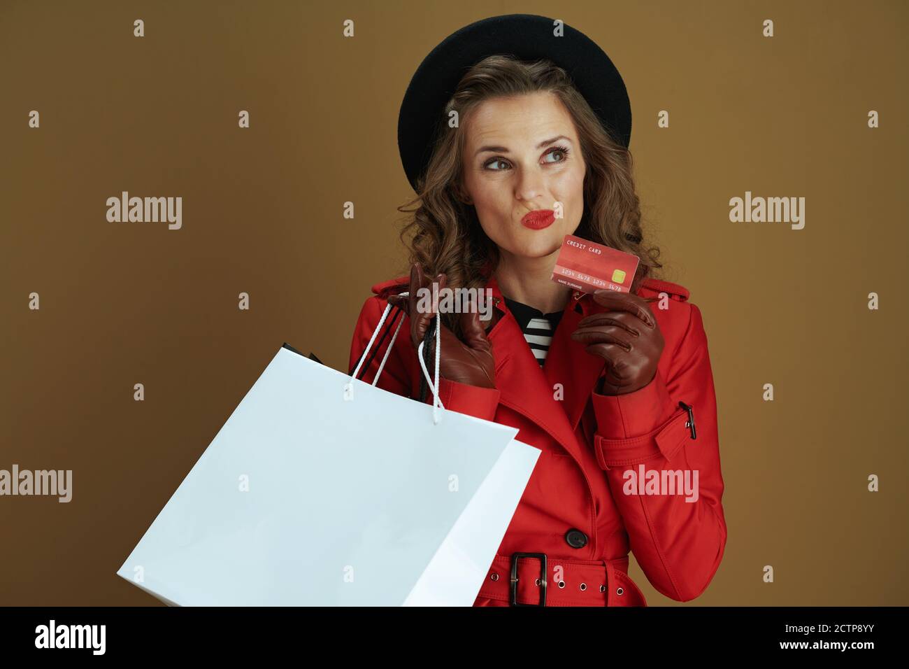 Hallo Herbst. Nachdenkliche trendige Hausfrau in rotem Mantel und schwarzer Baskenmütze mit Kreditkarte, Lederhandschuhen und Papier Einkaufstaschen vor Bronze Hintergrund. Stockfoto