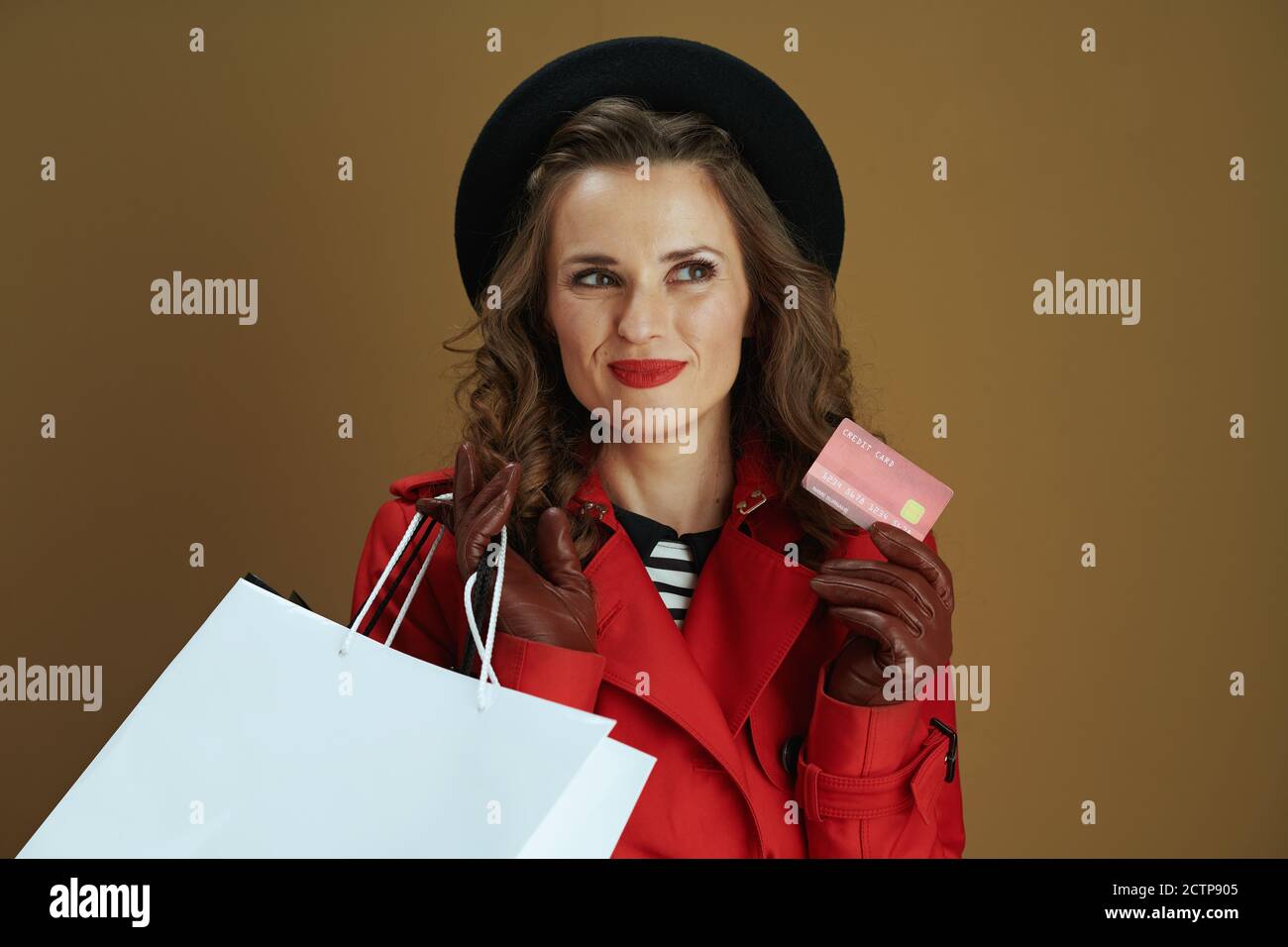 Hallo september. Nachdenkliche elegante 40 Jahre alte Hausfrau in rotem Mantel und schwarzer Baskenmütze mit Kreditkarte, Lederhandschuhe und Papier Einkaufstaschen isoliert Stockfoto