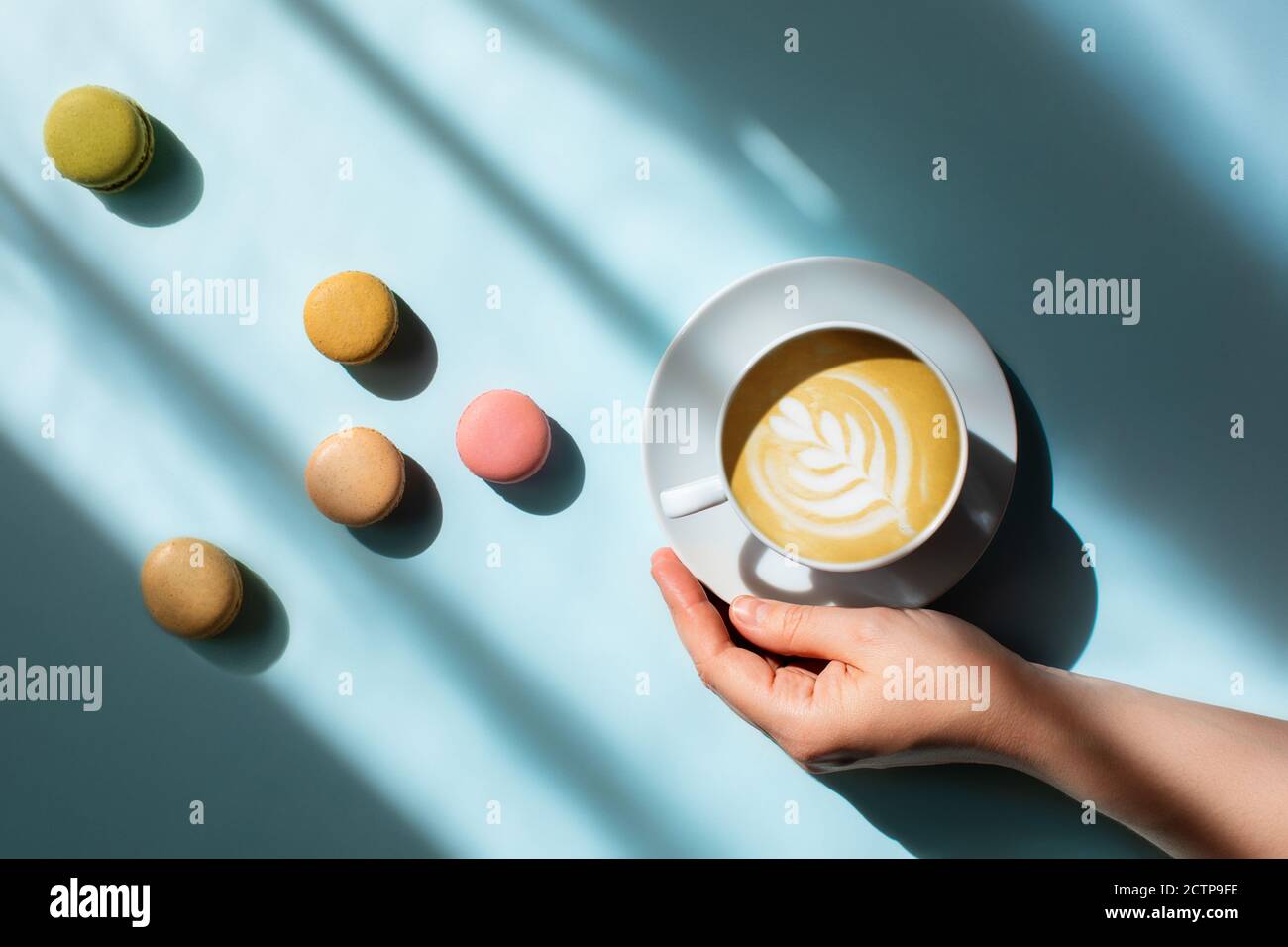 Morgenkaffee in der Hand einer Frau mit köstlichen Makronen, die im Sonnenlicht verstreut sind. Stockfoto