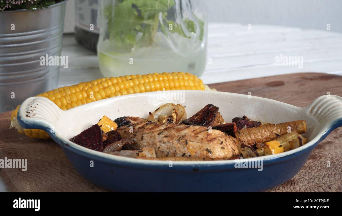 Ein gegrilltes Hähnchen sitzt auf einer Keramikplatte neben verschiedenen Wurzelgemüse. Verkohlte Linien aus dem Skillet-Muster sind deutlich sichtbar. Stockfoto