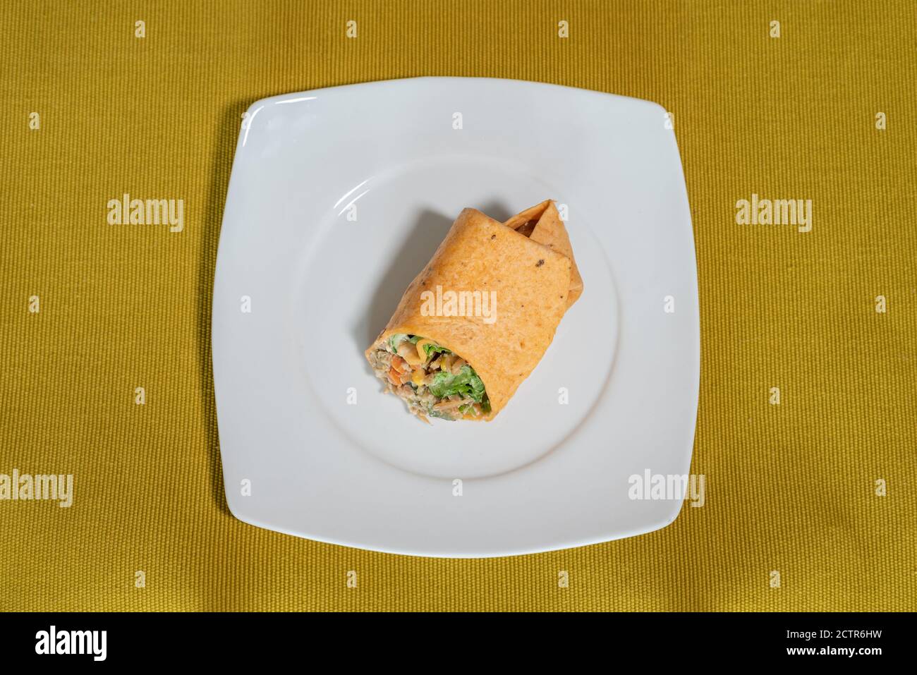 Vegetarische Verpackung serviert auf einem weißen Teller, der auf einem senffarbenen Tischläufer ruht, Food-Fotografie, die eine Tacos-Rolle zeigt, gefüllt mit Gemüse und Stockfoto