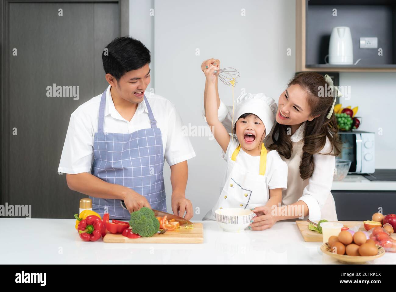 Asiatische Familie bricht ein Ei in die Schüssel und Tochter Rühreier in Schüssel auf weißem Tisch und sieht glücklich aus Essen kochen mit ihrem Vater und mot Stockfoto