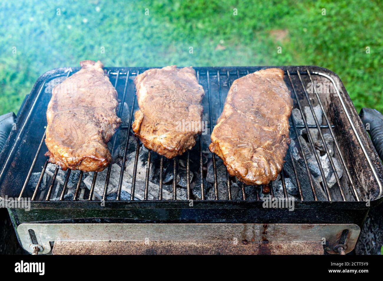 Nahaufnahme eines isolierten Bildes von drei Stücken mariniertem Flachsteak auf einem Holzkohlegrill. Der kleine zusammenklappbare Picknickgrill ist sehr heiß und Rauch kommt Stockfoto