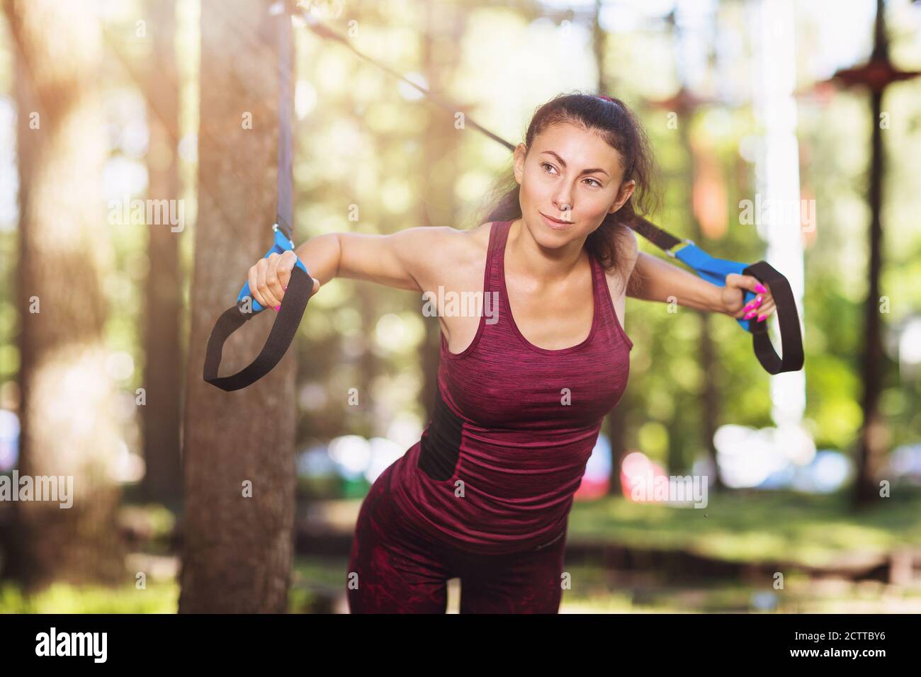Junge Frau macht Übungen im Park mit Fitness-Gurten an einem Baum befestigt. Ein Athlet trainiert seine Brustmuskeln an einem warmen Sommermorgen Stockfoto