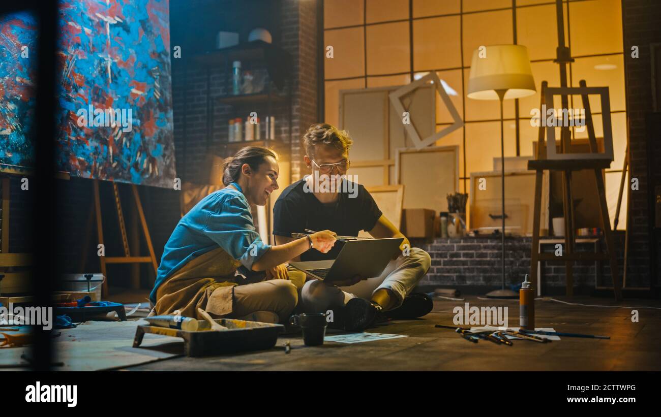 Einige Künstler sitzen in ihrem Studio verwenden Laptop-Computer, arbeiten an einem Projekt, reden und lächeln. Malerin und ihr Partner bei der Recherche Stockfoto