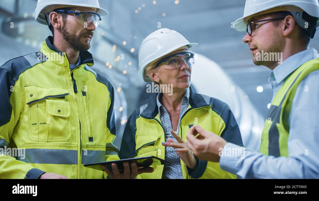 Drei Schwerindustrie-Ingenieure stehen in der Pipe Manufacturing Factory, verwenden digitale Tablet-Computer, haben Diskussion. Großes Rohr Montiert. Design und Stockfoto