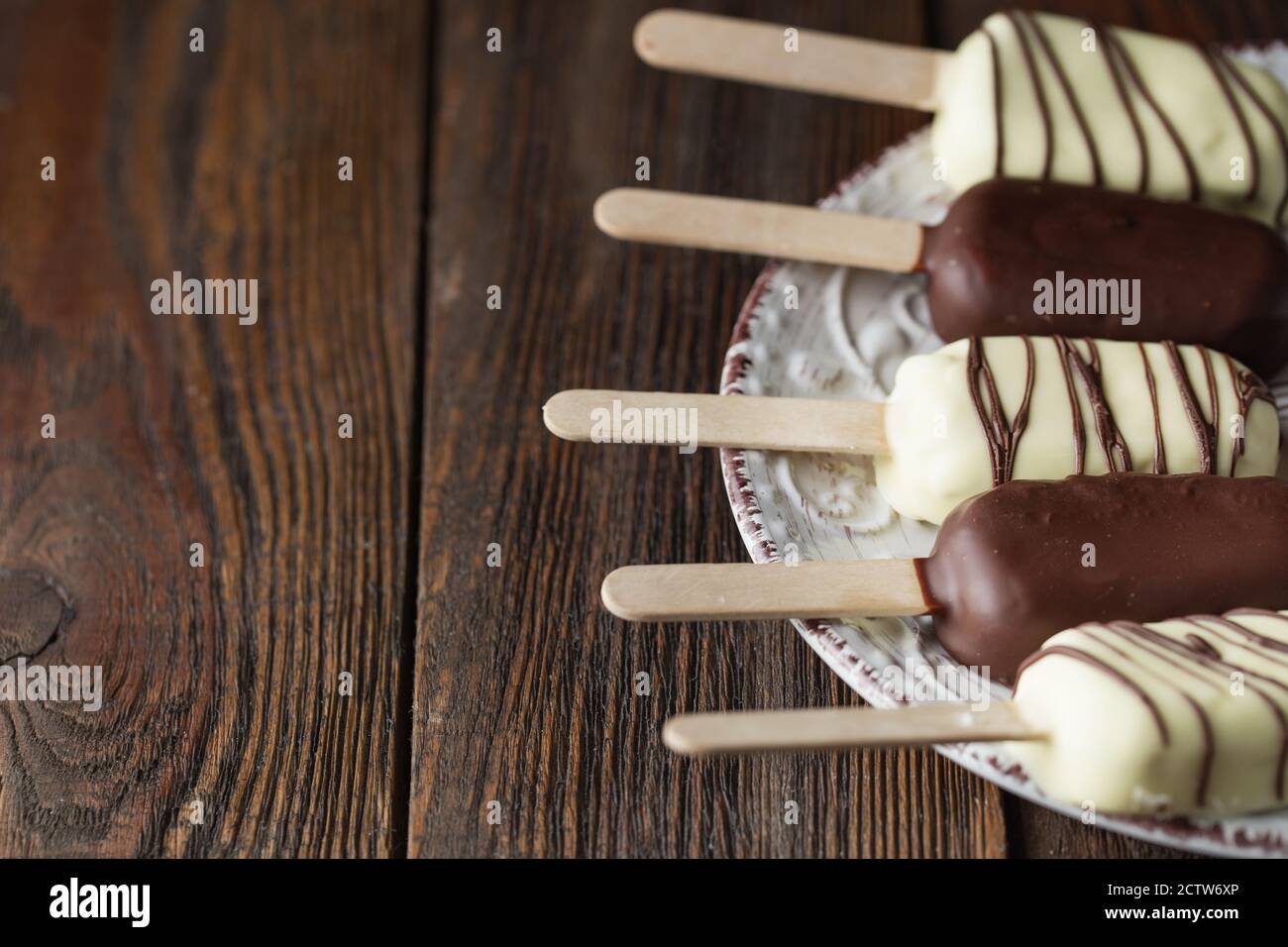 Milchschokolade-Eis auf einem Stock. Eisdielen mit weißer und dunkler Schokolade auf dem Teller auf Holzhintergrund bedeckt. Leerzeichen für Text. Choc Stockfoto