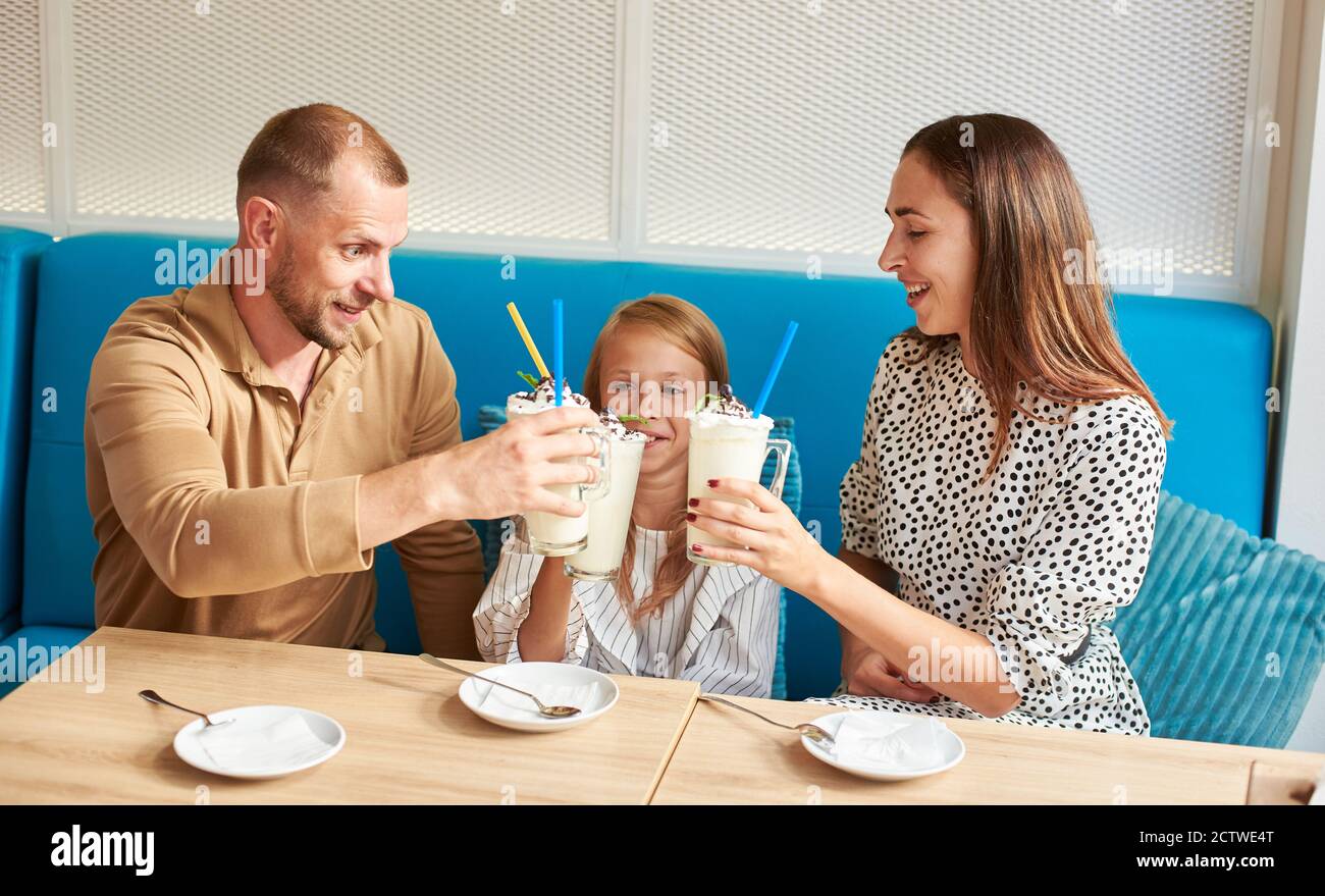 Glückliche Familie - Mama, Papa und Tochter sitzen im lokalen Café auf einem blauen Sofa und trinken Getränke, klirrende Gläser mit Milchshakes. Teller und Cocktaillöffel auf dem Tisch Stockfoto