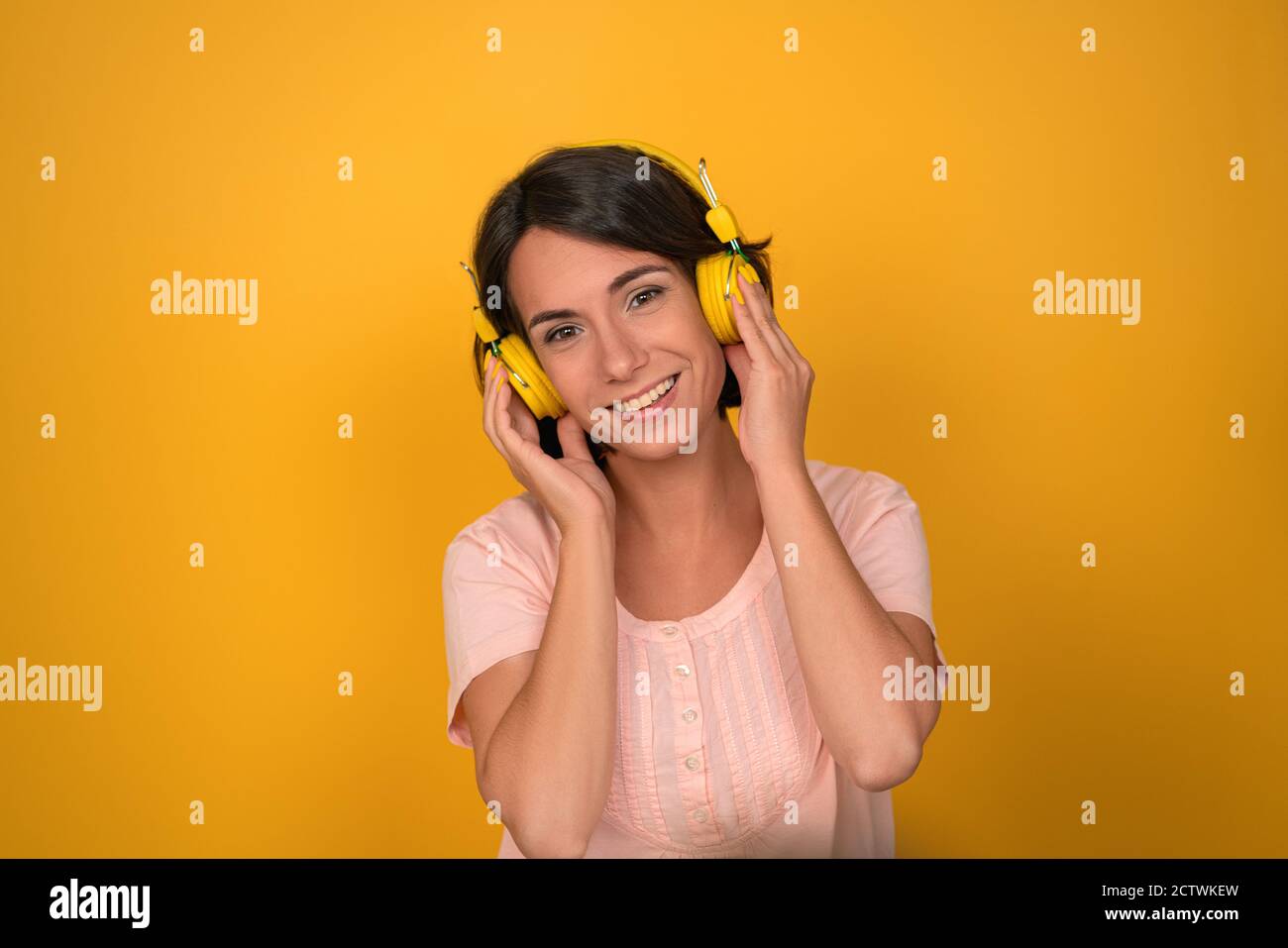 Die junge Frau drückt im Studio gelbe Kopfhörer an ihre Ohren. Brunette lächelt und hört Musik auf einem gelben Hintergrund. Hochwertige Fotos Stockfoto