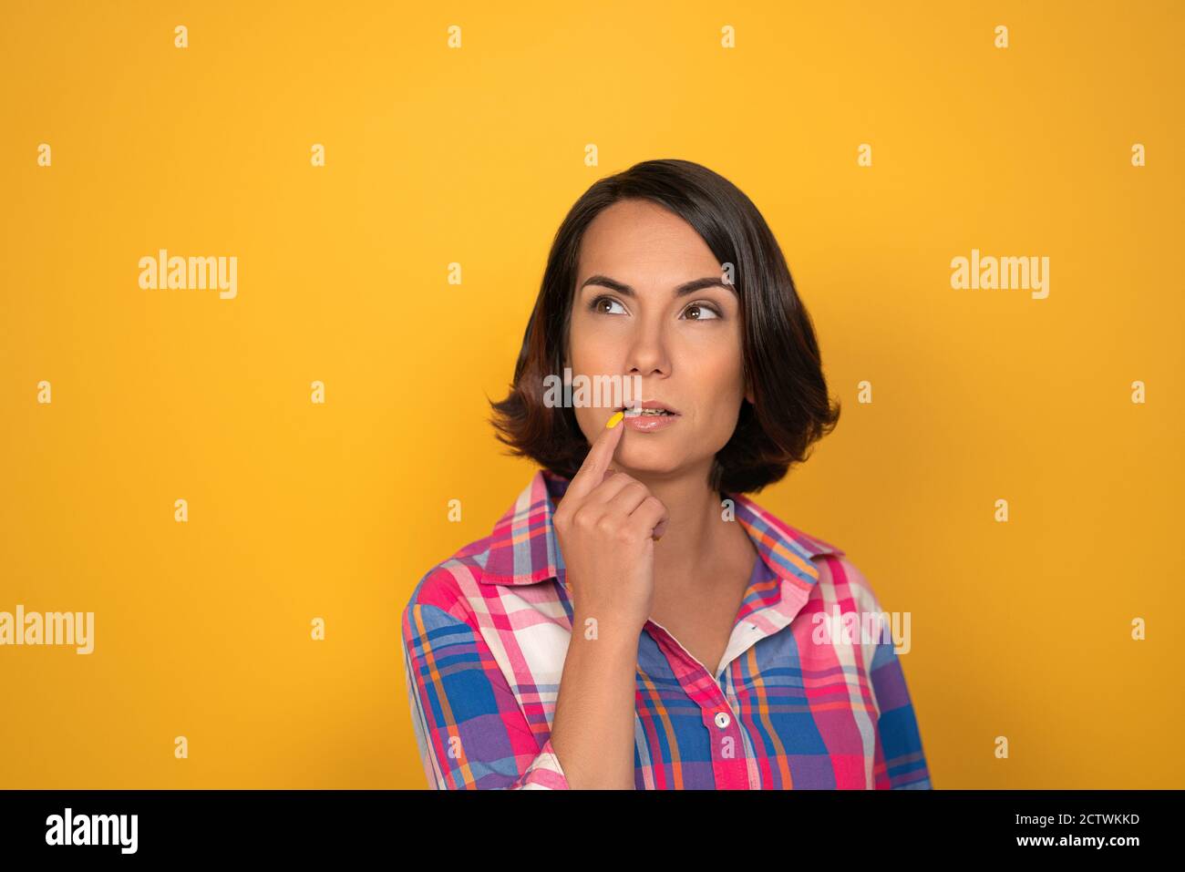 Brunette Frau macht einen brütenden Blick auf einem gelben Hintergrund im Studio. Frau legt ihren Finger auf ihre Lippen und schaut mit einer gewölbten Augenbraue nach oben Stockfoto