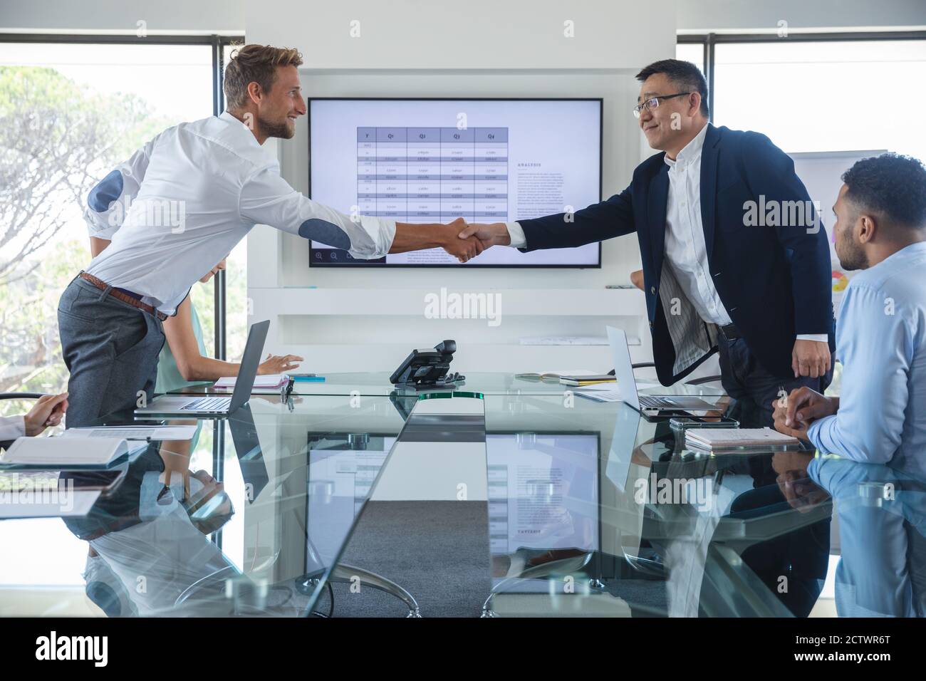 Asiatischer und kaukasischer Geschäftsmann, der bei einem Geschäftstreffen die Hände über den Tisch schüttelt und Kollegen neben ihnen sitzen. Kreatives Geschäft Stockfoto