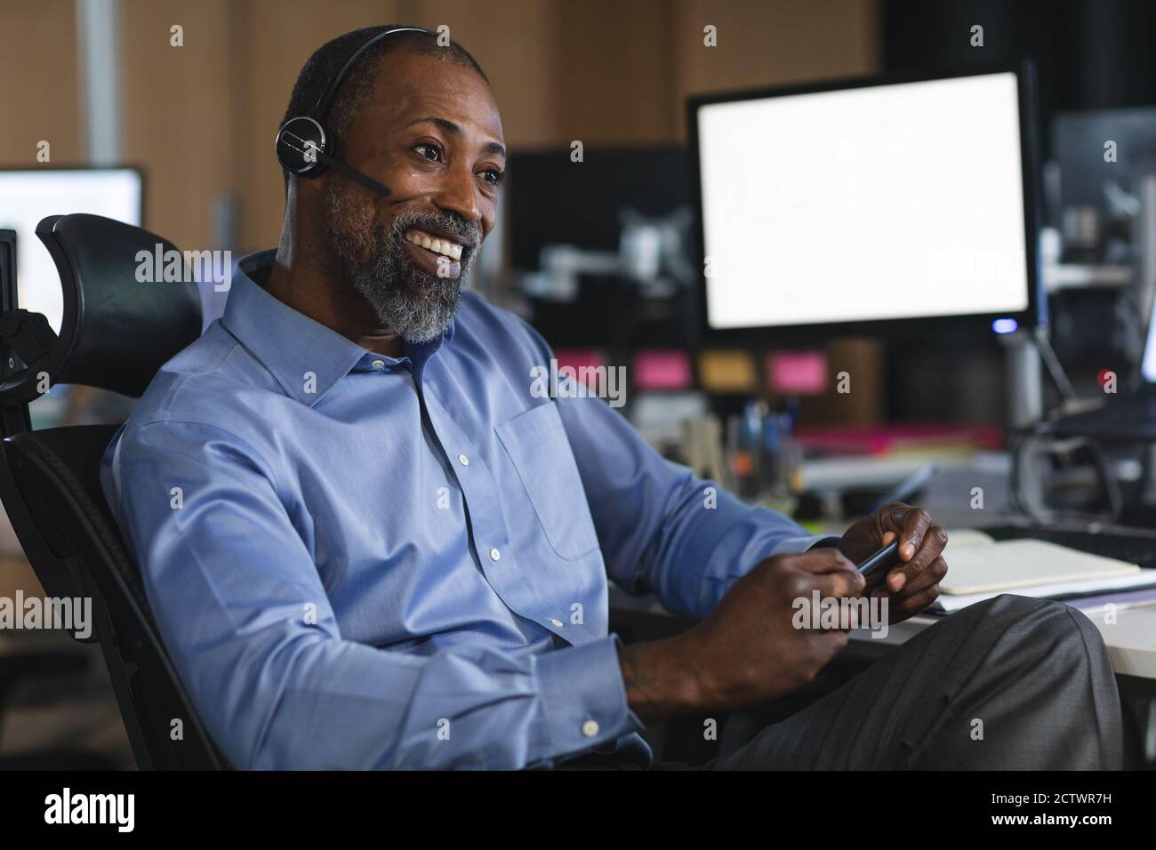 Afroamerikanischer Geschäftsmann, der spät abends in einem modernen Büro arbeitet, an einem Schreibtisch sitzt, ein Headset trägt und lächelt. Stockfoto