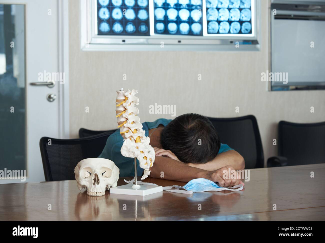 Ein asiatischer Neurochirurg fühlt sich müde und schläft in der medizinischen Praxis. Schädel- und Lendenwirbelsäule Modell auf dem Tisch, Gehirn-MRT an Bord. Stockfoto