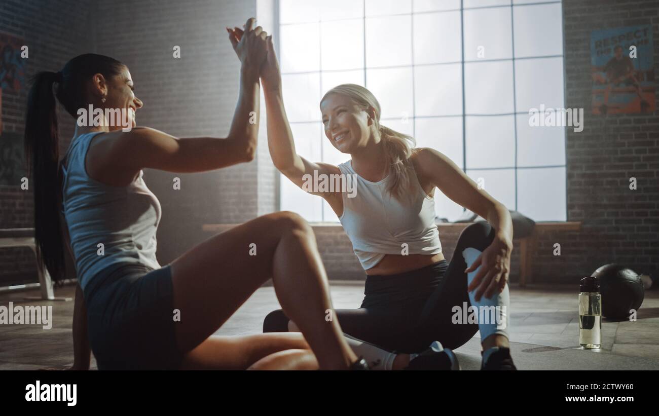 Zwei schöne Fit Athletic Mädchen sitzen auf einem Boden der Industrial Loft Gym. Sie sind mit ihrem Trainingsprogramm zufrieden und geben erfolgreich einen High Five. Stockfoto