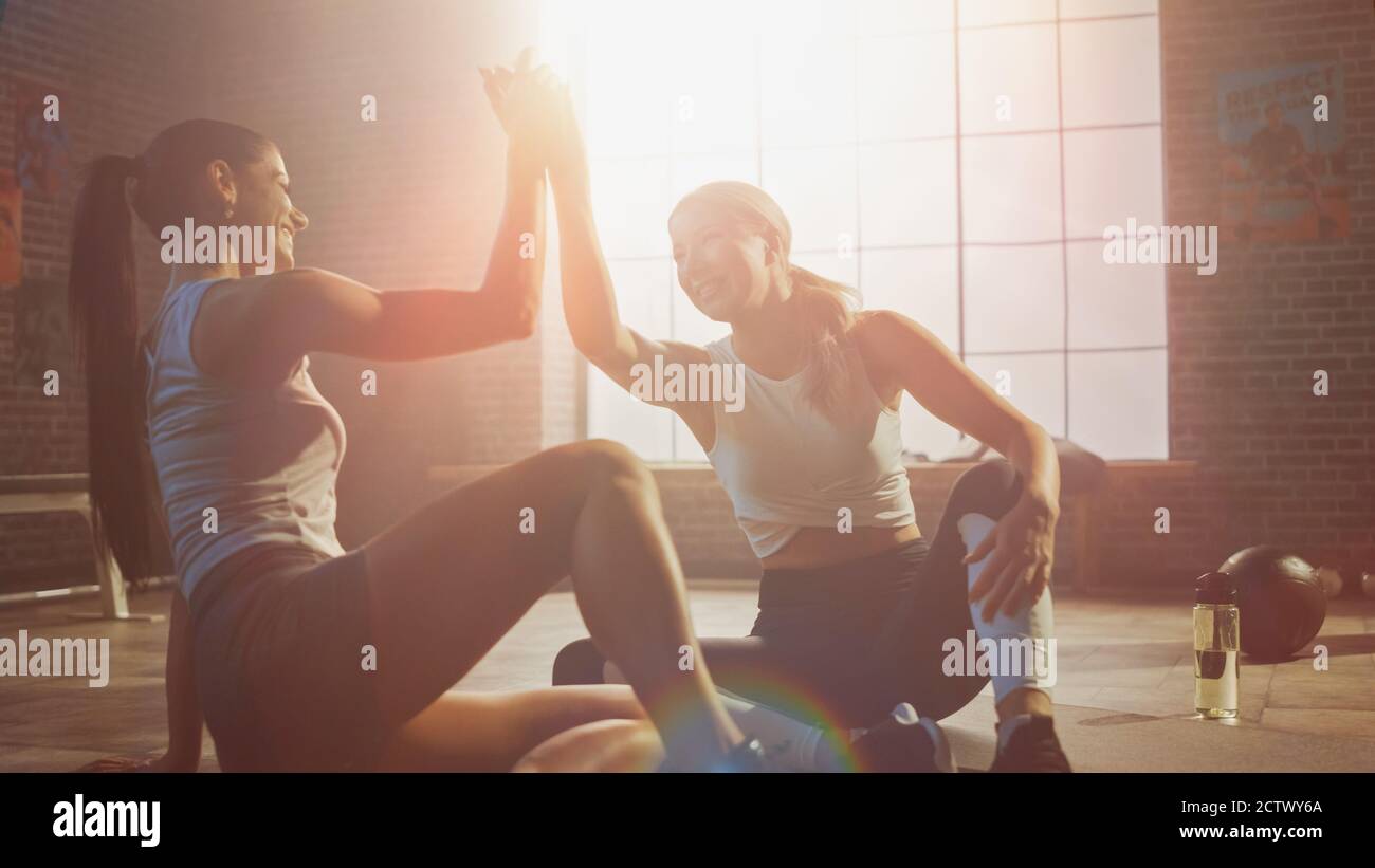 Zwei schöne Fit Athletic Mädchen sitzen auf einem Boden der Industrial Loft Gym. Sie sind mit ihrem Trainingsprogramm zufrieden und geben erfolgreich einen High Five Stockfoto
