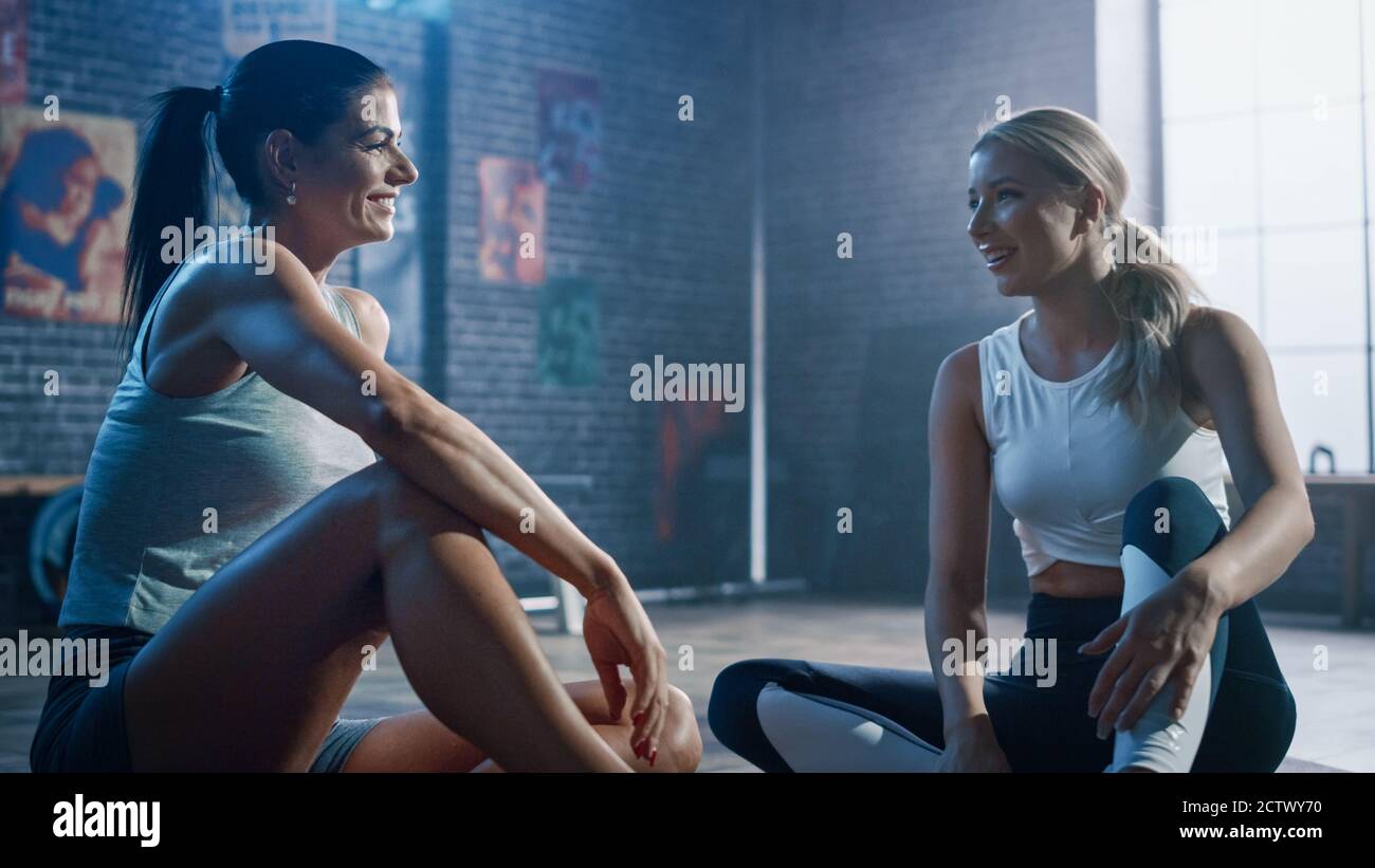 Zwei schöne Fit Athletic Mädchen sitzen auf einem Boden der Industrial Loft Gym. Sie sind mit ihrem Trainingsprogramm zufrieden und sprechen über sportliche Erfolge Stockfoto