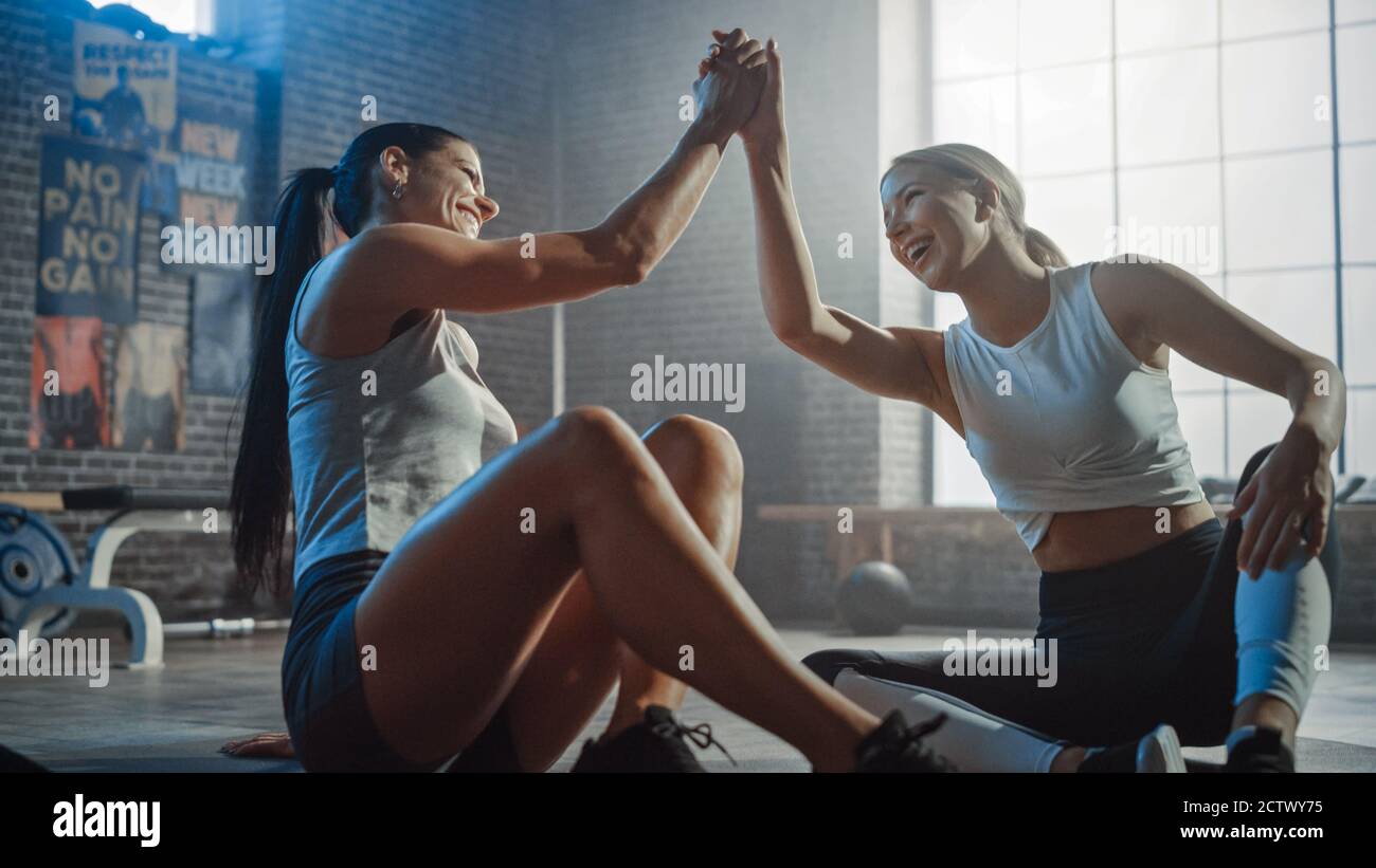 Zwei schöne Fit Athletic Mädchen sitzen auf einem Boden der Industrial Loft Gym. Sie sind mit ihrem Trainingsprogramm zufrieden und geben erfolgreich einen High Five. Stockfoto