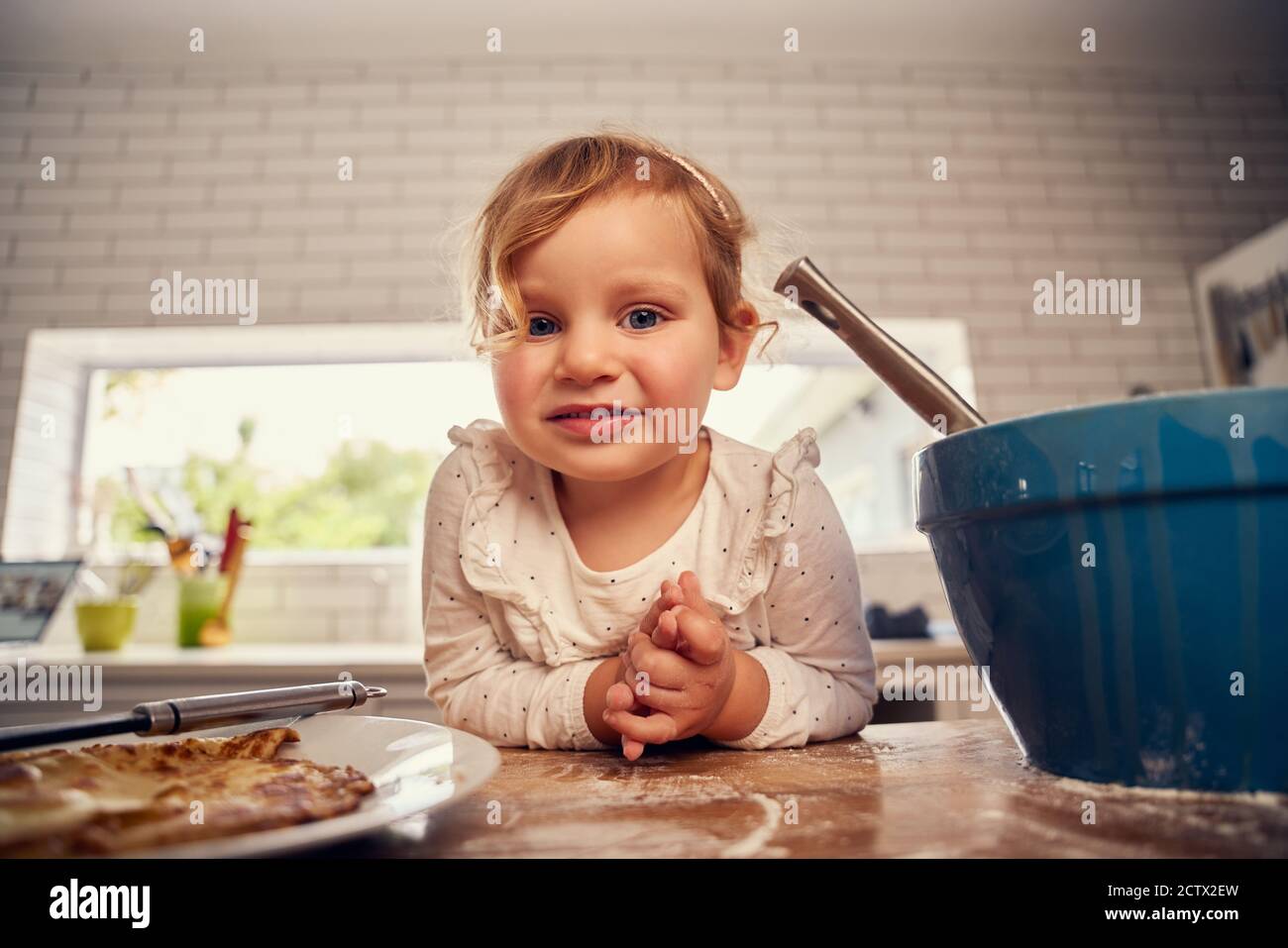 Portrait von kleinen niedlichen Mädchen lehnte sich auf Küche Zähler während Kochen mit Utensilien und lächeln und Blick auf die Kamera Stockfoto
