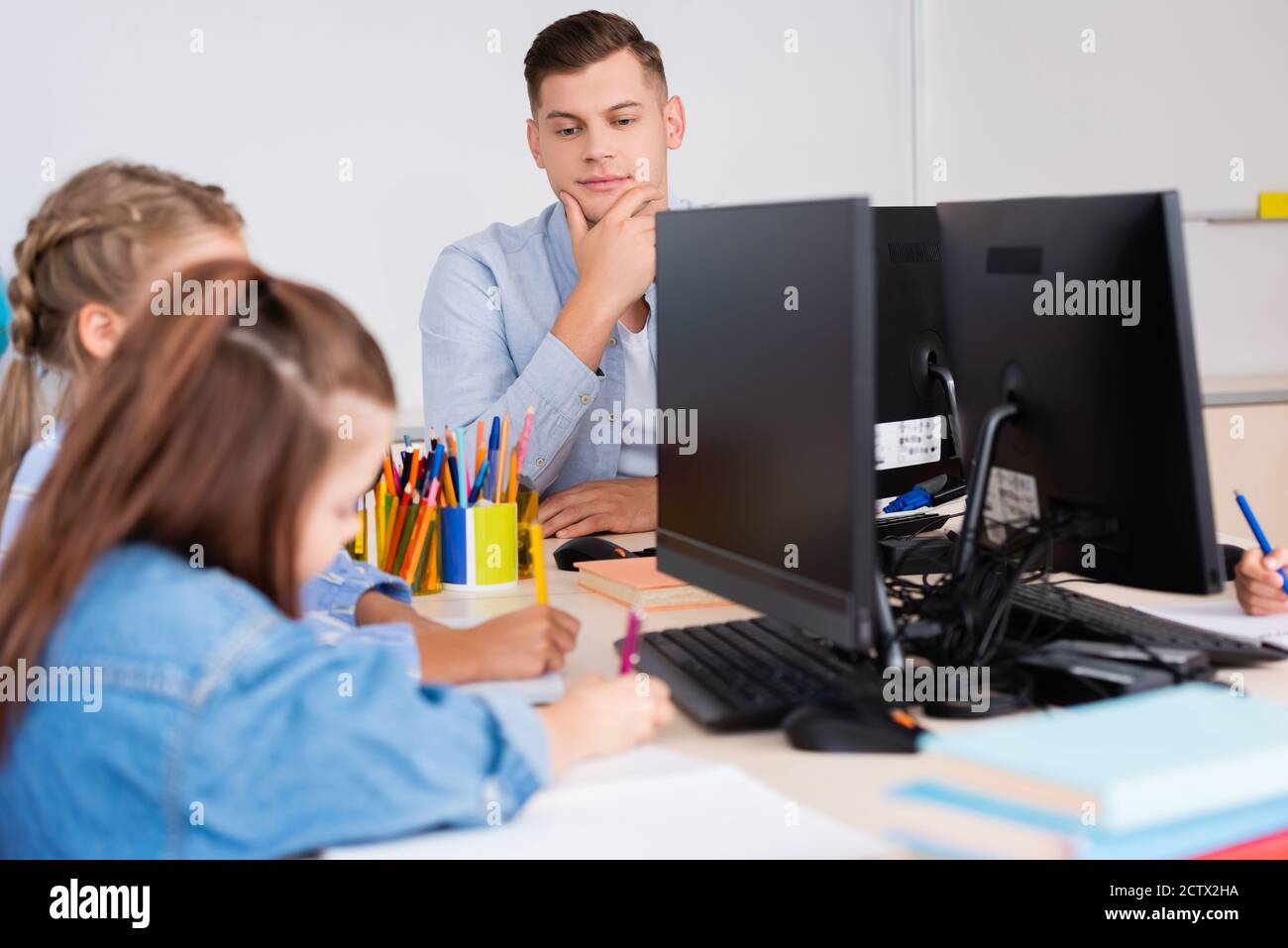 Selektiver Fokus des Lehrers, der in der Nähe der Schüler sitzt und auf Notizbüchern schreibt In der Nähe von Computern in der Schule Stockfoto