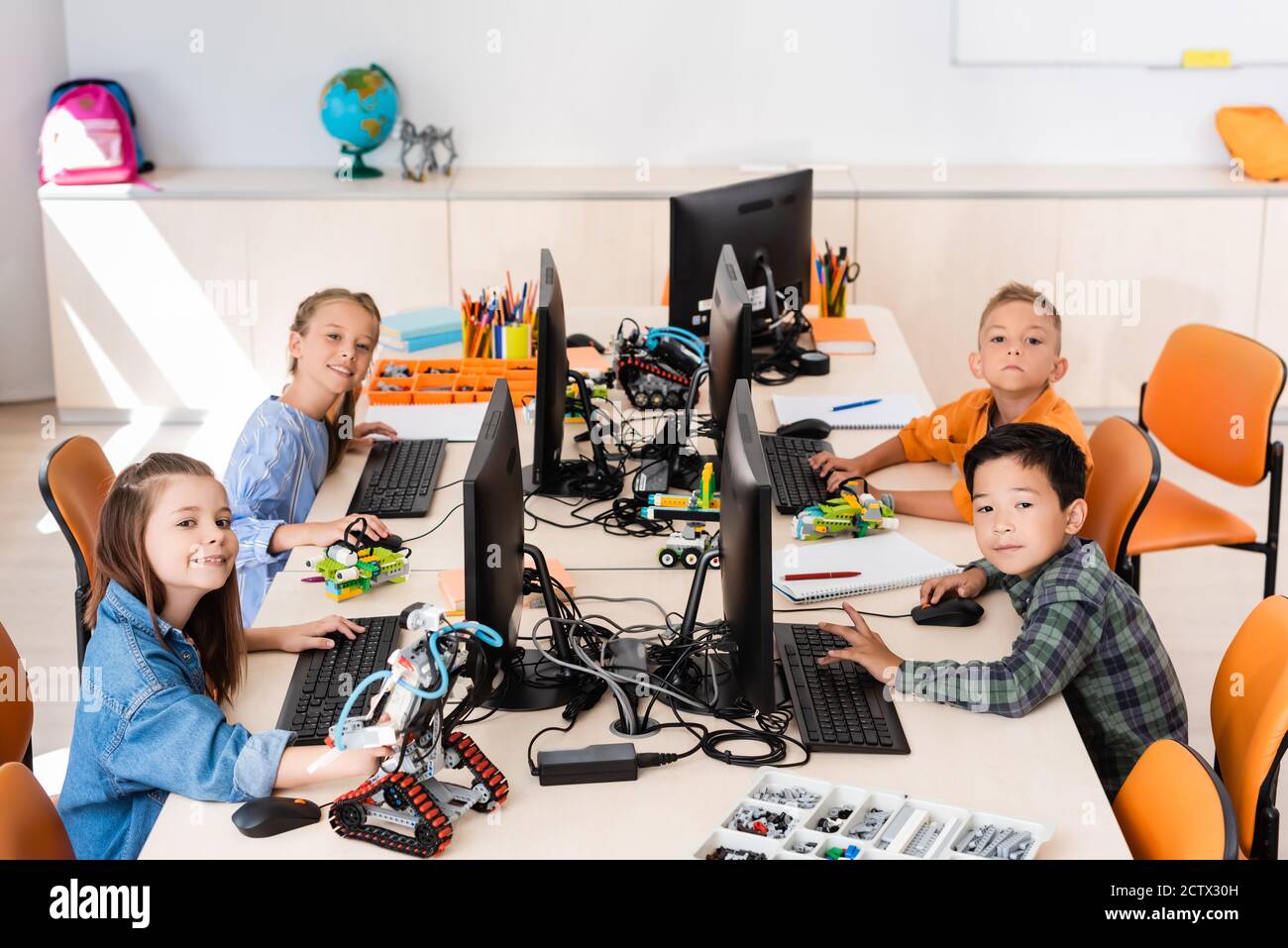 Selektiver Fokus von multikulturellen Schulkindern, die in der Nähe von Robotern auf die Kamera schauen Und Computer im Klassenzimmer Stockfoto