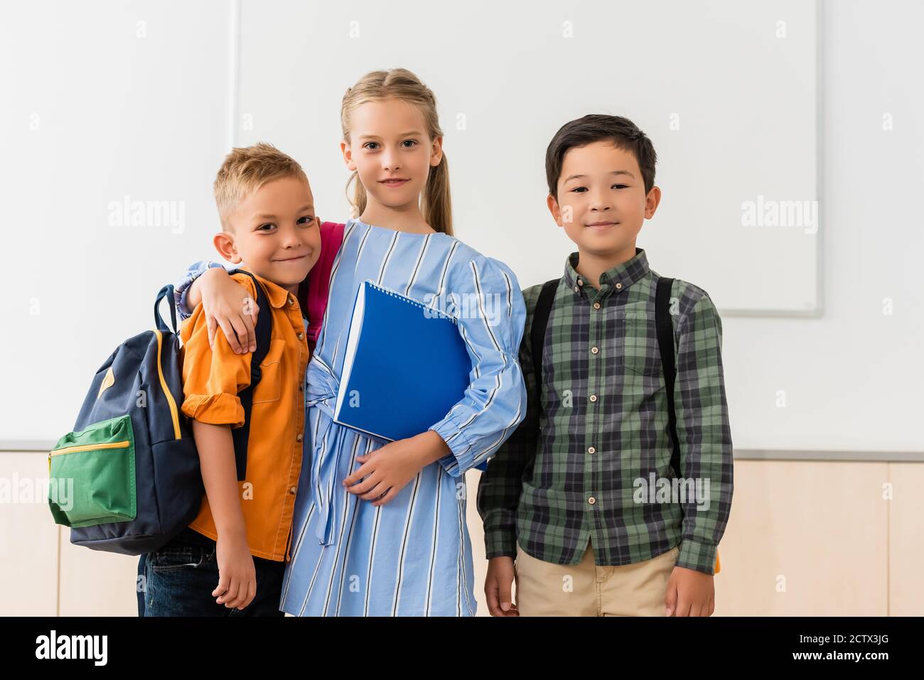 Schulmädchen mit Notebook umarmt Klassenkamerad in der Nähe asiatische Freund im Klassenzimmer Stockfoto