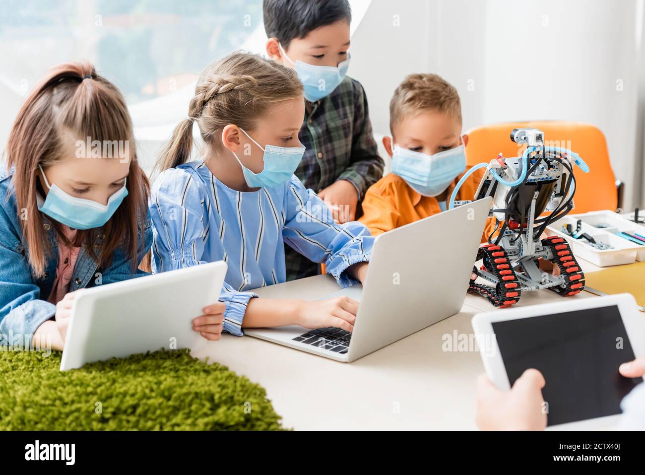 Selektive Fokussierung multiethnischer Kinder in medizinischen Masken mit digitalen Tablet und Laptop in der Nähe Lehrer und Roboter in der Schule Stockfoto