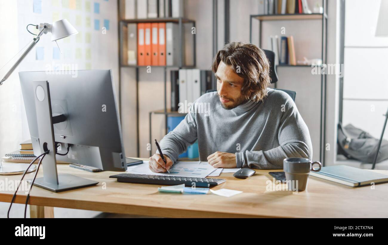 Gutaussehende langhaarige Unternehmer sitzen an seinem Schreibtisch im Büro arbeitet auf Desktop-Computer, Arbeiten mit Dokumenten, Korrigieren von Diagrammen, Zeichnen Stockfoto
